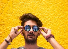 Young man with sunglasses standing against a vibrant yellow wall in daylight.